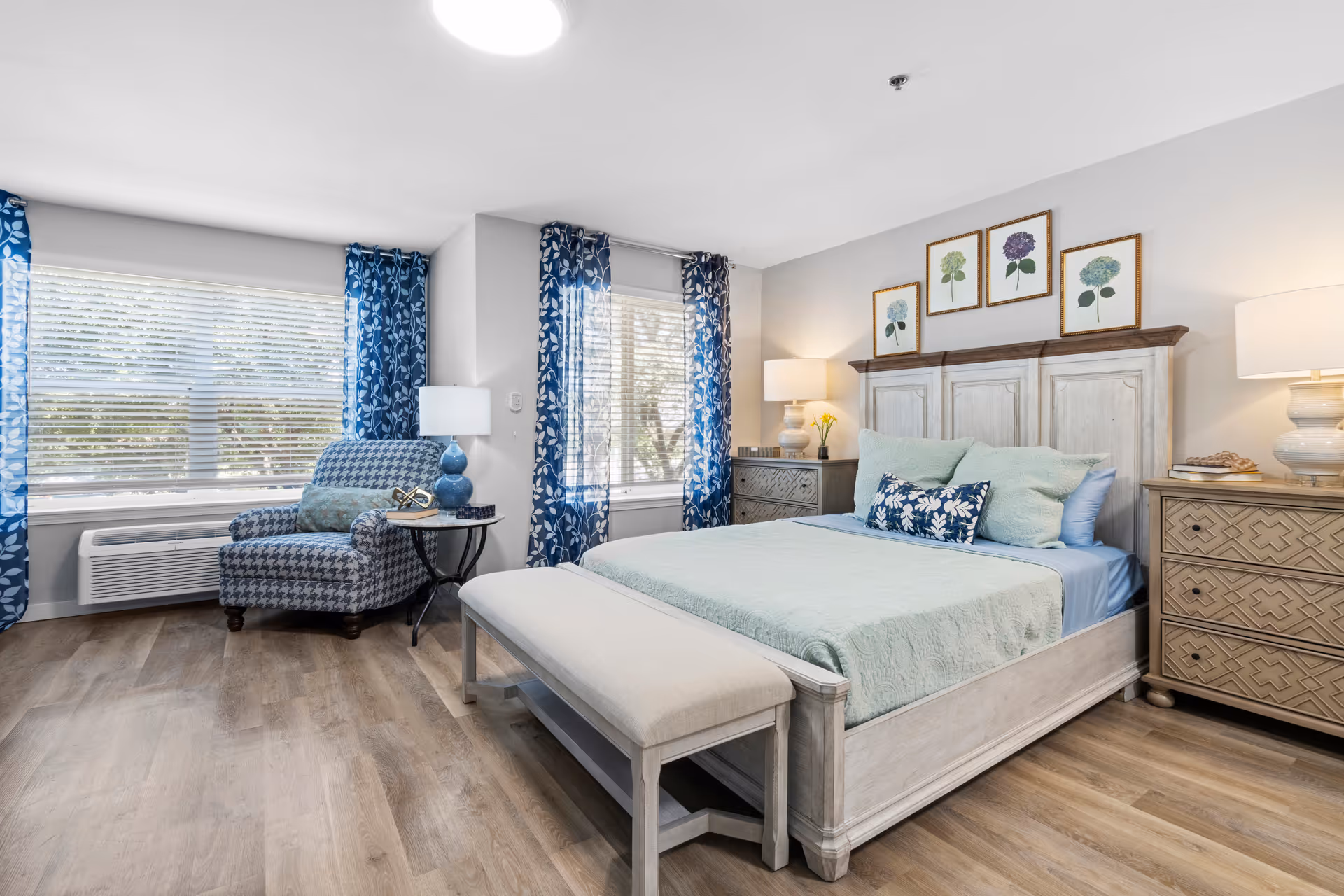 A bright and airy bedroom featuring a large bed with light blue and green bedding, a cushioned bench at the foot of the bed, two matching nightstands with lamps, and a patterned armchair near two windows with blue floral curtains. The walls are decorated with framed botanical prints, and the floor has a wood finish.
