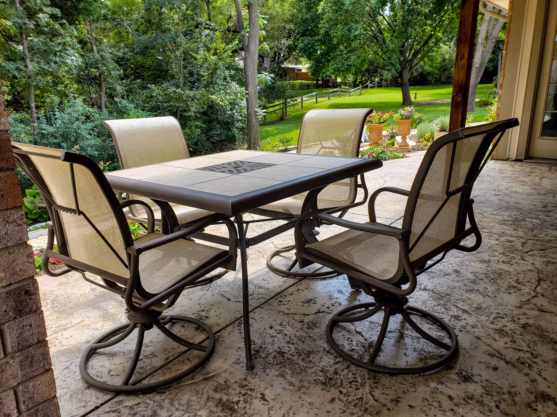 Outdoor patio area with a square table and four mesh chairs on a textured concrete floor, surrounded by greenery and trees in the background.