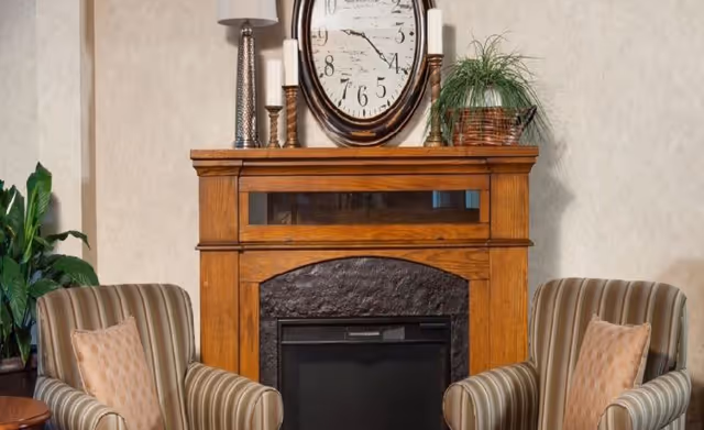 A cozy seating area with two striped armchairs flanking a wooden fireplace mantel topped with a round clock and decorative items.