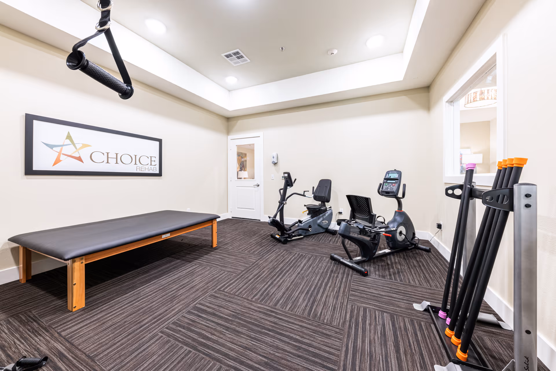 Small rehab/fitness room with a therapy table, two recumbent exercise bikes, and a rack of fitness poles under recessed lighting.