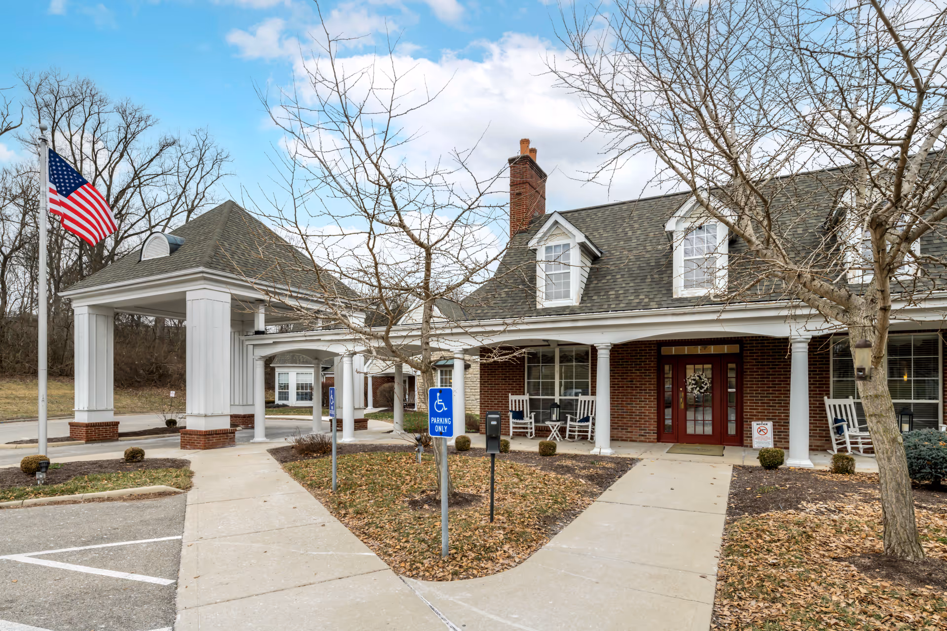 Exterior view of Brookdale Englewood facility showing a covered entrance with white columns, a brick building with dormer windows, an American flag on a flagpole, leafless trees, and a sidewalk leading to the entrance. There are rocking chairs on the porch and handicap parking signs near the entrance.