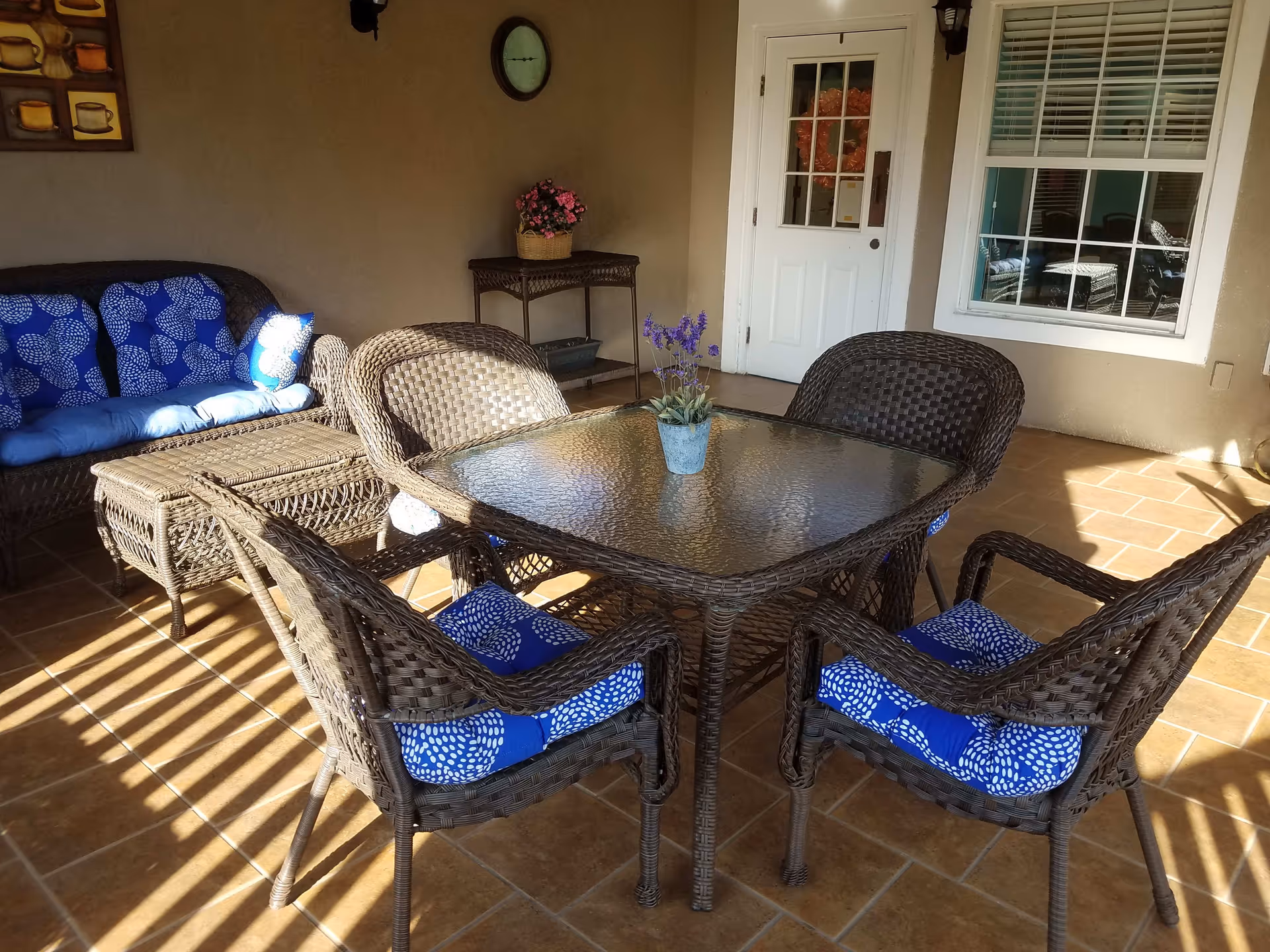 Wicker patio dining set with a glass-top table and blue-cushioned chairs on a tiled covered porch next to a sofa and potted plant.