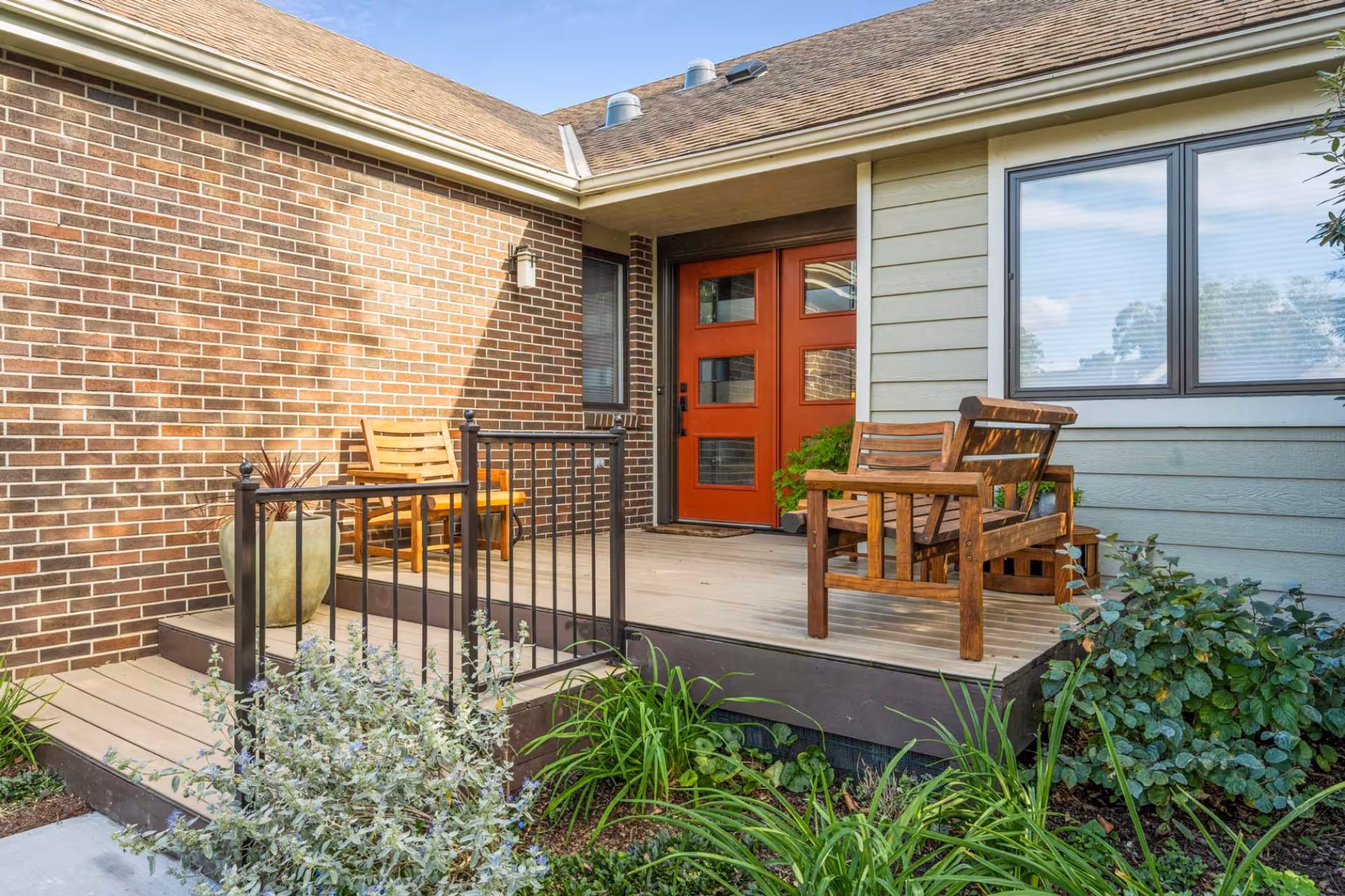 Outdoor patio area with wooden chairs and a small table, adjacent to a brick and siding building with a red door and a window. There are plants and greenery around the patio.