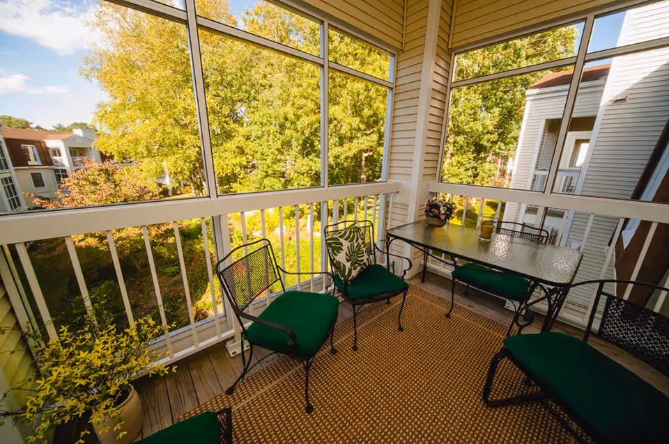 A screened-in balcony with a glass-top table and four metal chairs with green cushions. There is a potted plant on the floor and a small flower arrangement on the table. The balcony overlooks a green, leafy outdoor area with trees and neighboring buildings.