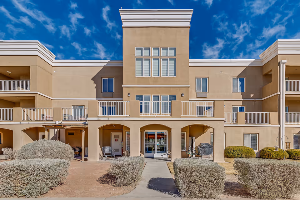 Exterior view of a beige multi-story senior living facility building with balconies, arched entryways, and trimmed bushes in front under a blue sky with scattered clouds.