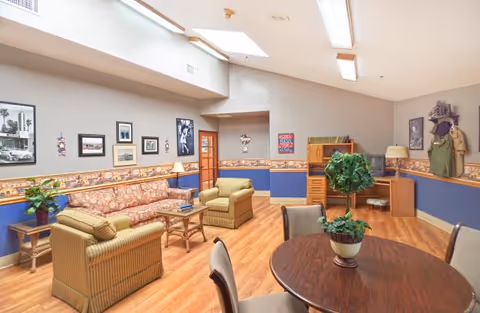 A cozy living room area in a senior living facility with wooden flooring, a patterned sofa, two armchairs, a round wooden table with chairs, and a potted plant on the table. The walls are decorated with framed pictures and a wallpaper border, and there is a wooden desk with a lamp and a TV in the corner.