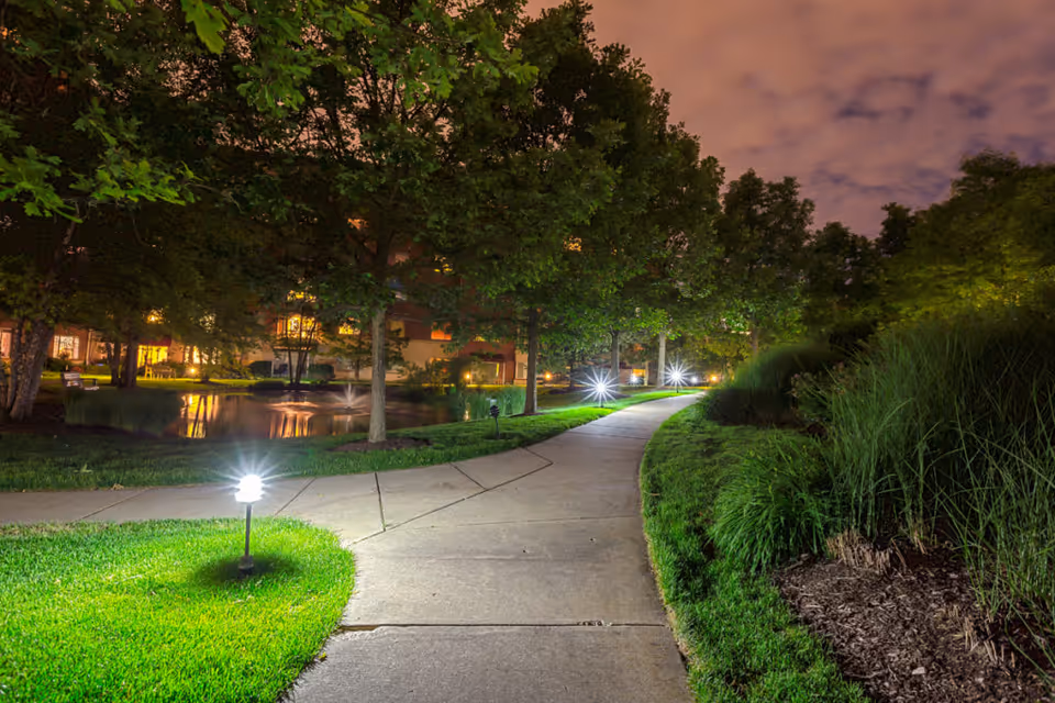 Nighttime illuminated walkway through landscaped grounds with trees, a pond, and a lit building in the background.