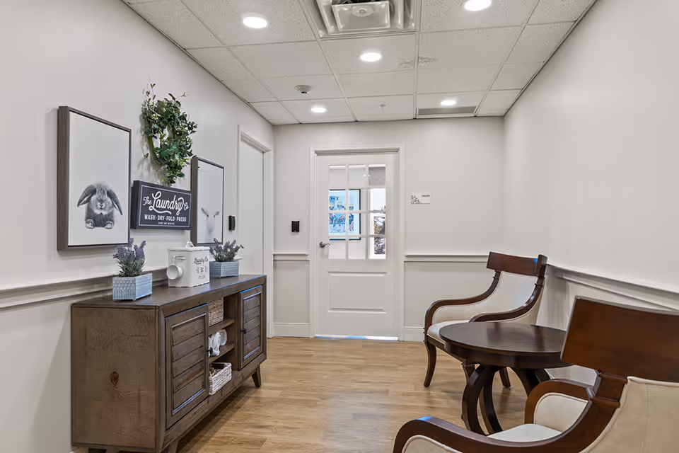 A clean, well-lit hallway with light-colored walls and wooden flooring. On the left side, there is a wooden cabinet decorated with small potted plants, a tissue box, and framed pictures including a rabbit and a sign that reads 'The Laundry Wash - Dry - Fold - Press'. On the right side, there are two wooden chairs with light cushions and a round wooden table between them. At the end of the hallway, there is a white door with glass panels.
