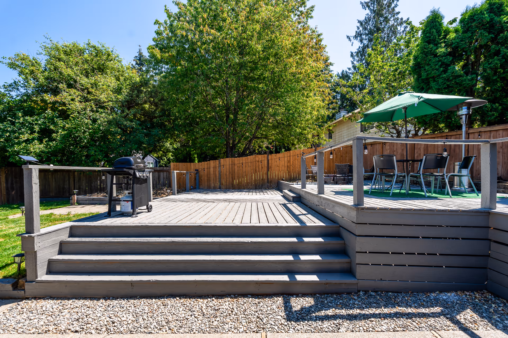 Outdoor wooden deck area with stairs leading up to it, featuring a barbecue grill on the left side and a seating area with a table, chairs, and a green umbrella on the right. The deck is surrounded by a wooden fence and lush green trees under a clear blue sky.