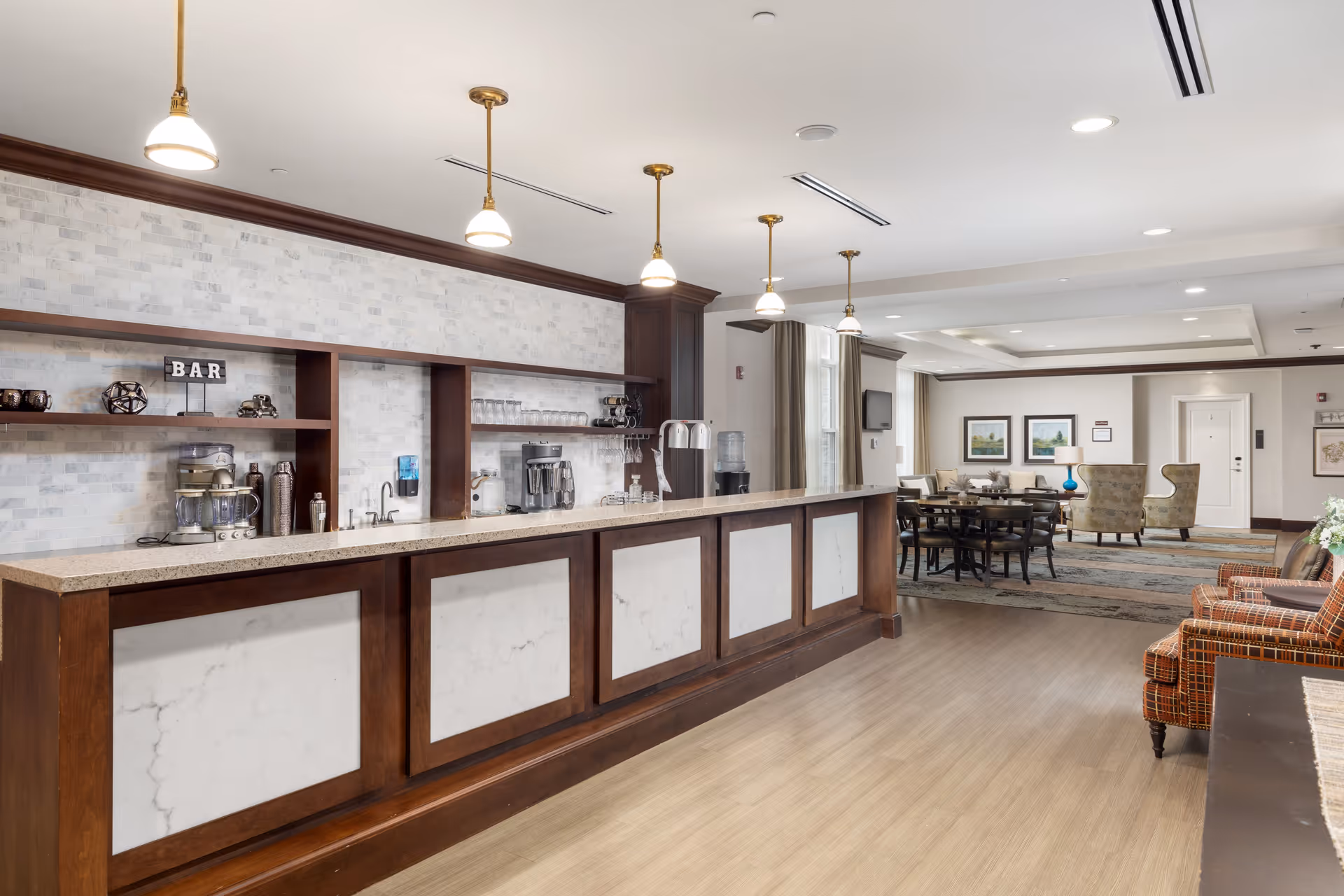A spacious and well-lit common area in an assisted living facility featuring a long counter with a marble-like front and wooden trim, pendant lights hanging above, and shelves behind the counter with coffee machines, cups, and a small sign that says 'BAR'. In the background, there are tables and chairs arranged for dining or socializing, along with comfortable armchairs and framed artwork on the walls.