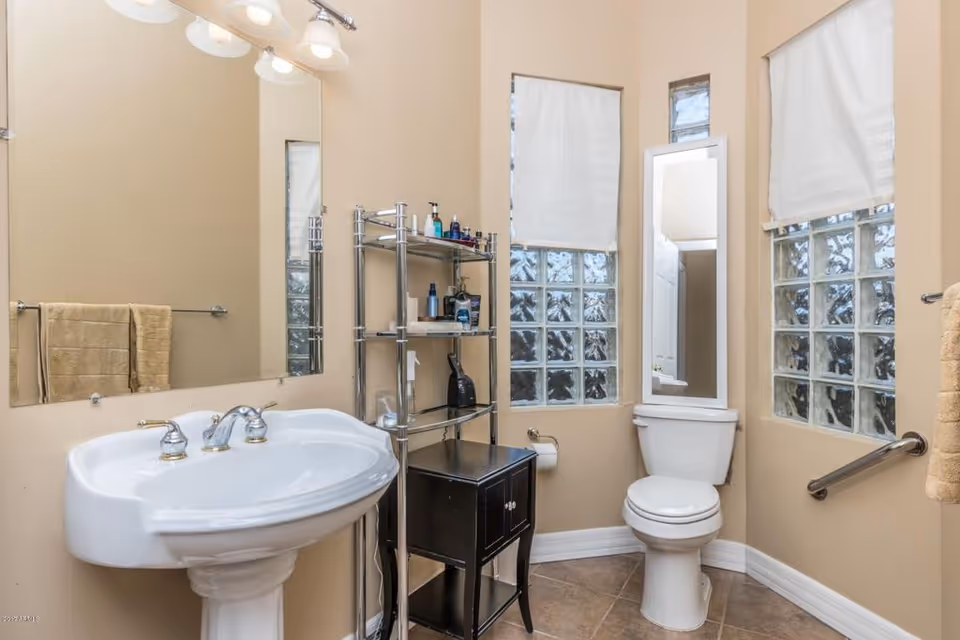 A bathroom with beige walls and tiled floor featuring a white pedestal sink with silver faucets, a large mirror above the sink, a towel rack with beige towels, a metal shelving unit with toiletries, a small black cabinet, a white toilet with a tall mirror behind it, and two windows with frosted glass blocks and white blinds.