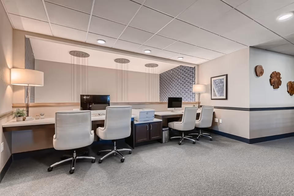 A modern office or computer workstation area with four white swivel chairs positioned at a long desk. The desk has two desktop computers, a printer, and two table lamps. The room features neutral-colored walls, carpeted floor, and decorative wall art including framed pictures and wooden wall hangings.