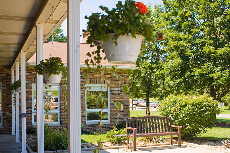 Outdoor view of a senior living facility with a covered walkway supported by white columns, hanging flower pots with green plants and red flowers, a wooden bench on a paved area, green bushes, trees, and a brick building in the background under a clear blue sky.