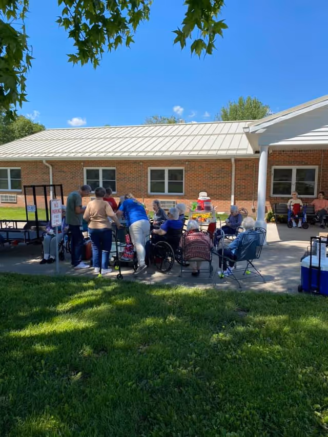 A group of elderly people, some in wheelchairs, gathered outside on a patio area next to a brick building under a clear blue sky. Several caregivers or visitors are standing and interacting with the seated individuals. There is green grass in the foreground and some leafy tree branches hanging from the top of the image.