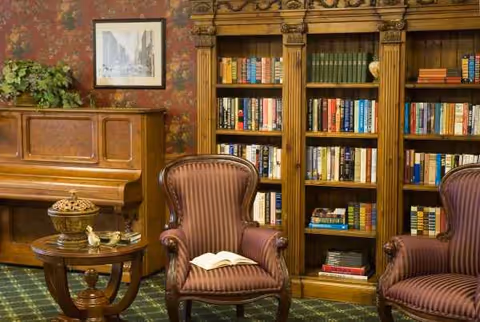 A cozy senior living lounge with antique piano, tall bookshelf, and striped armchairs.