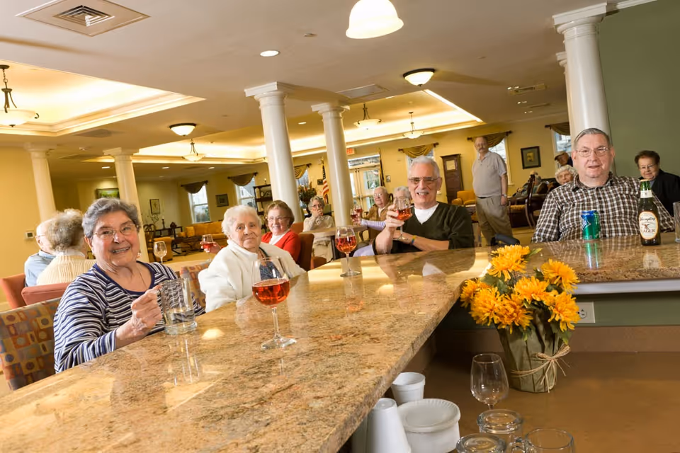 A group of elderly people sitting around a large granite countertop in a well-lit common area with columns and ceiling lights. Some are holding drinks including wine and beer, and there is a vase with yellow flowers on the counter. The room has a warm and inviting atmosphere with comfortable seating and windows in the background.