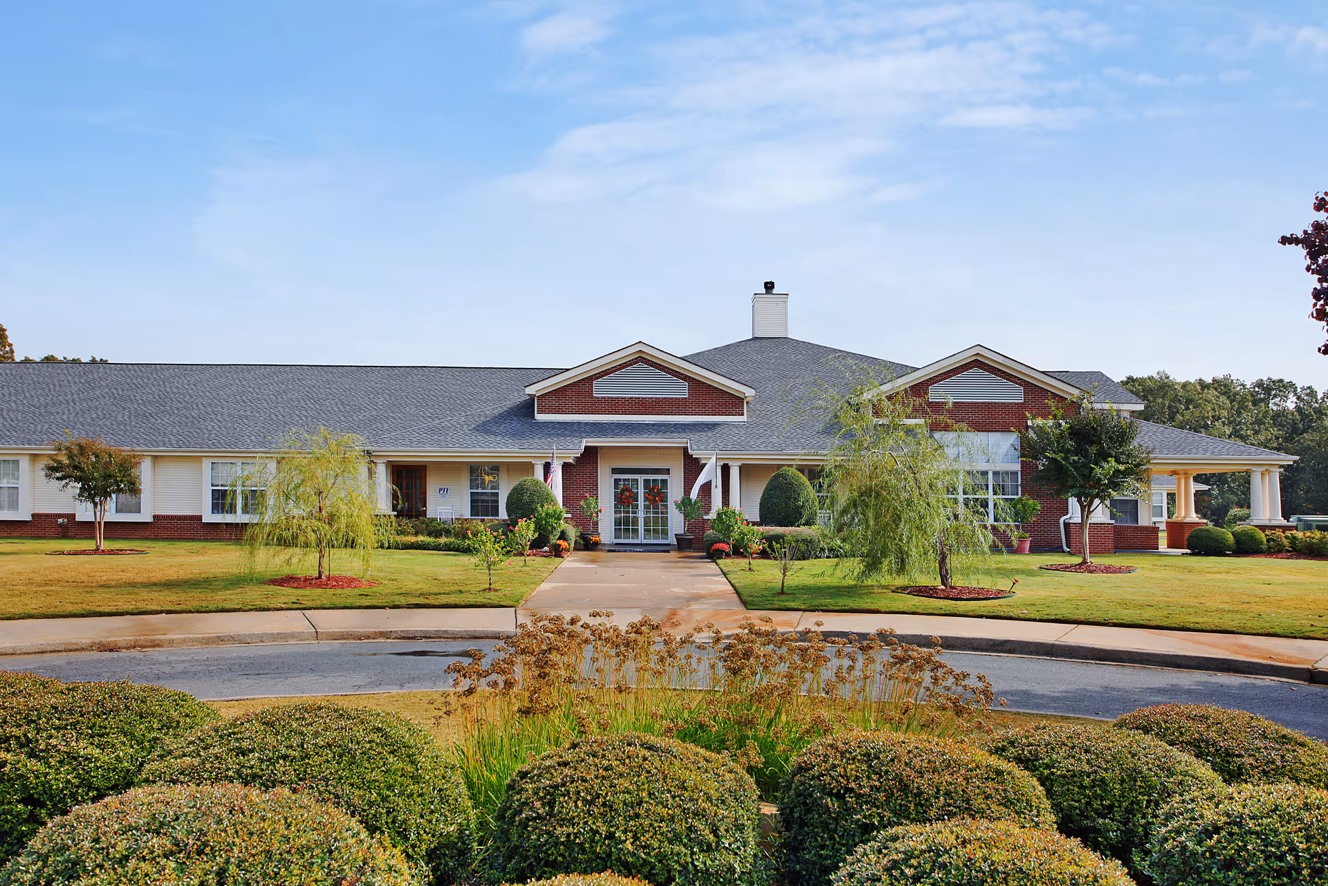 Front exterior view of Willow Grove of Sherwood, a single-story building with a gray roof and red brick accents. The entrance is centered with a walkway leading to double glass doors, surrounded by neatly trimmed bushes and small trees on a well-maintained lawn under a clear blue sky.