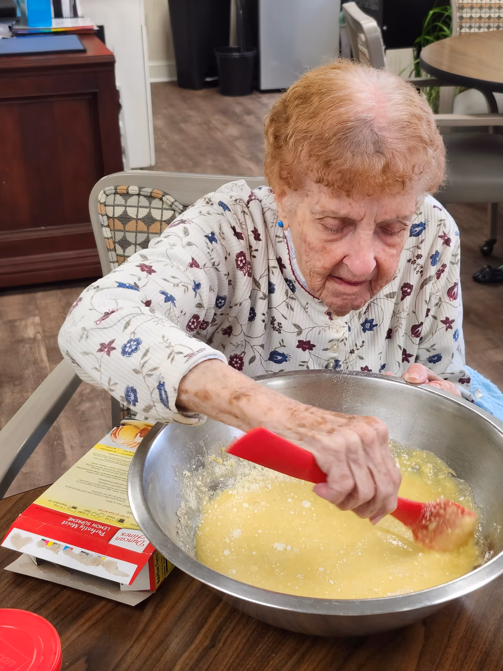 An elderly woman with short curly hair wearing a white floral patterned shirt is sitting at a table and stirring a large metal mixing bowl with a red spatula. There is a box of Duncan Hines lemon supreme cake mix on the table beside her. The setting appears to be an indoor common area with wooden flooring and chairs.