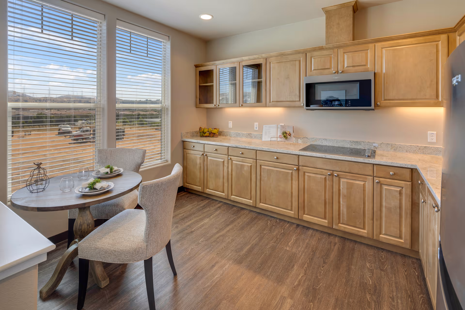 A bright kitchen with light wood cabinets, a built-in microwave, and a smooth cooktop. There is a small round dining table with two upholstered chairs near large windows with blinds, offering a view of an outdoor area with vehicles and hills in the distance. The floor is a dark wood finish.