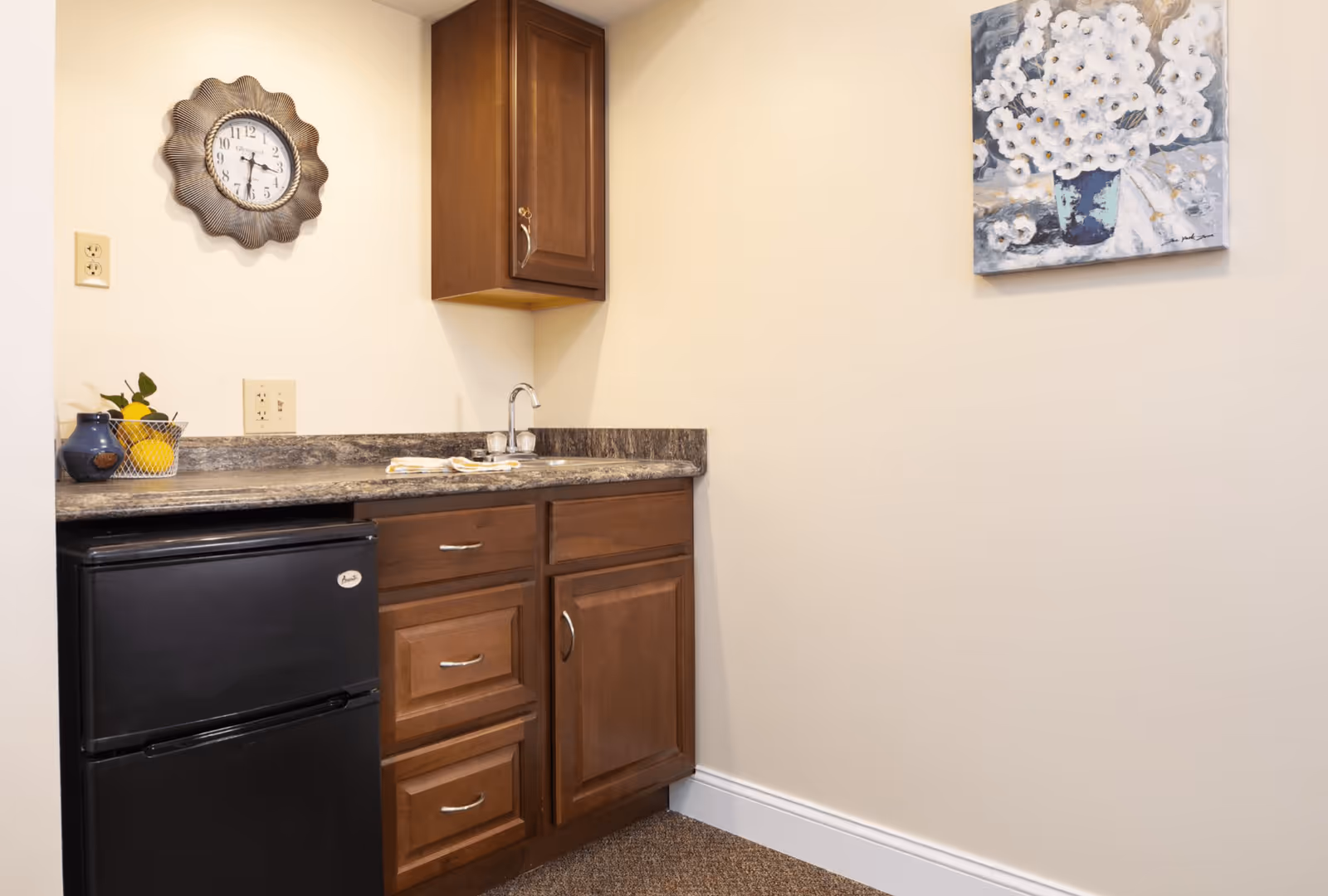 Small kitchenette with dark wood cabinets, a mini fridge, countertop sink, wall clock and a floral painting on a cream wall.