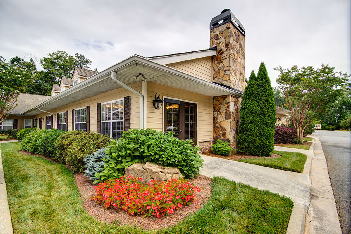 Exterior view of a single-story building with beige siding, multiple windows with dark shutters, a stone chimney, and a well-maintained garden with green shrubs and red flowers along a concrete walkway.