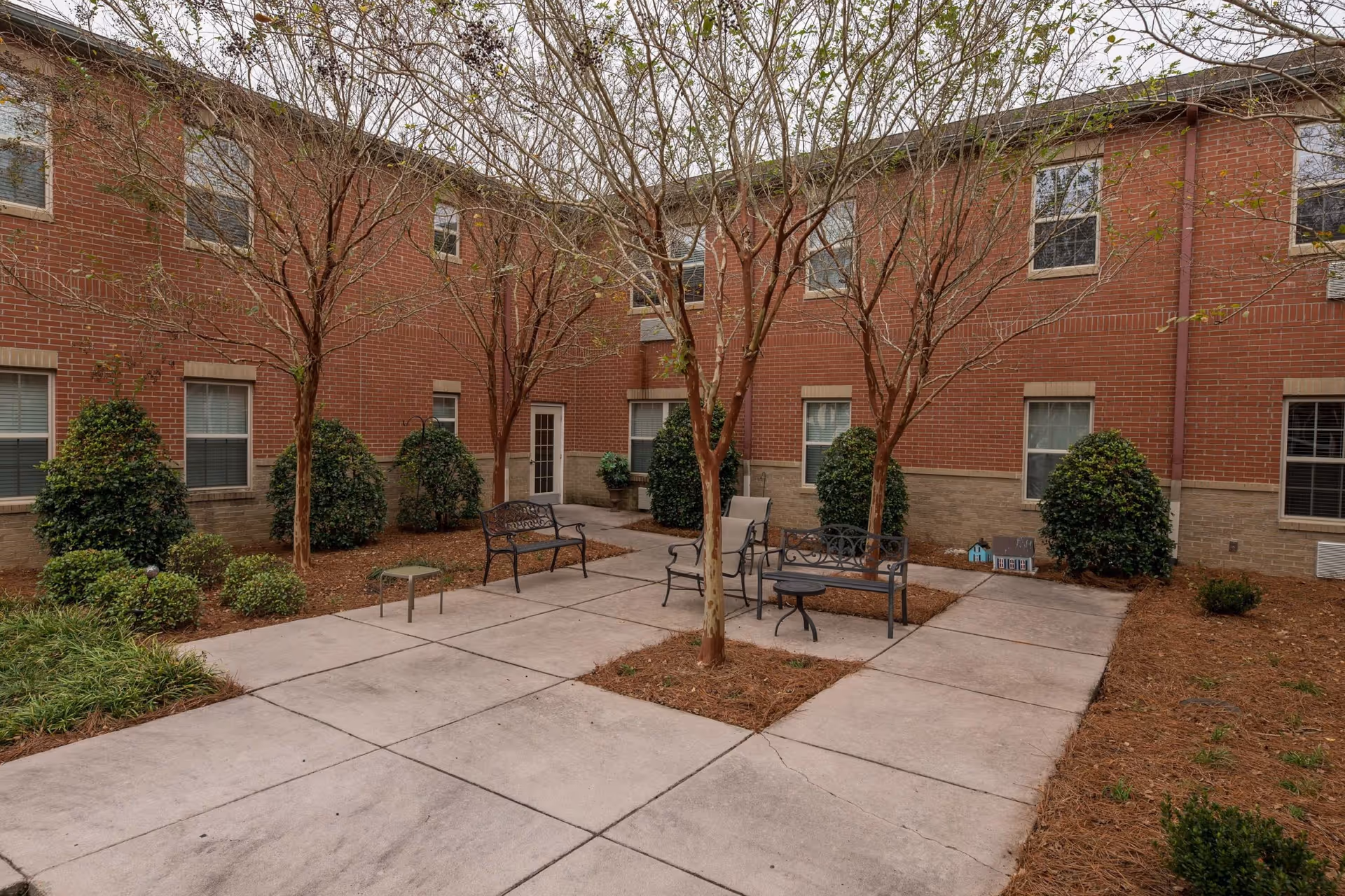 Outdoor courtyard area with brick building walls surrounding it. The courtyard has several leafless trees, neatly trimmed bushes, and paved walkways. There are metal benches and chairs arranged around the courtyard for seating.