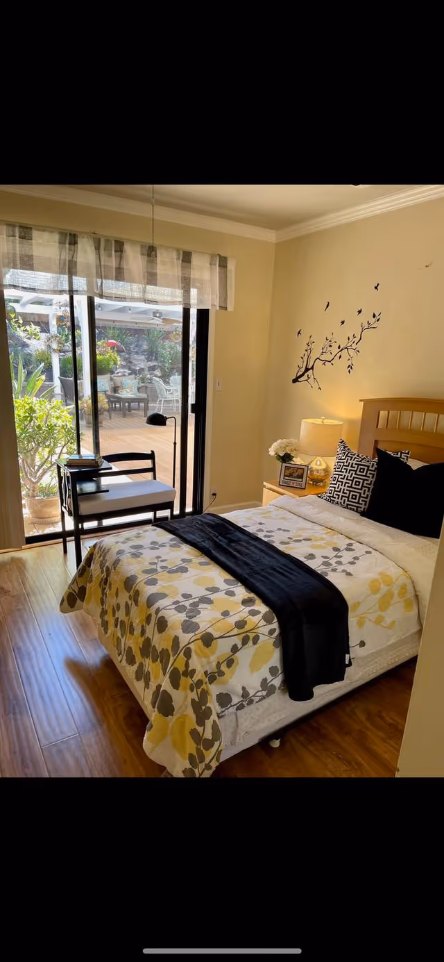 A cozy bedroom with a bed covered in a floral patterned bedspread in yellow, gray, and white. The bed has black and patterned pillows and a black throw blanket. Next to the bed is a nightstand with a lamp, a small flower arrangement, and a framed photo. On the wall above the nightstand is a decorative wall decal of a tree branch with birds. A sliding glass door with sheer curtains leads to an outdoor patio area with plants and seating visible outside. The room has wooden flooring and light-colored walls.