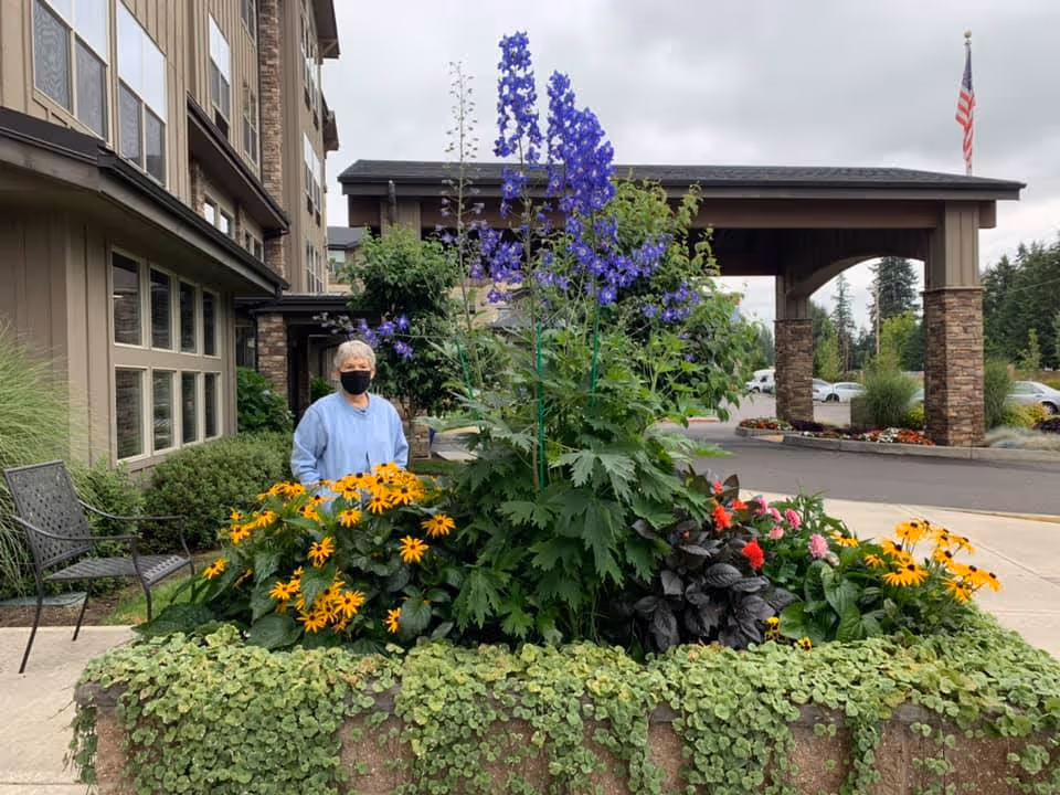 An elderly person wearing a light blue jacket and a black face mask stands behind a large flower bed filled with yellow, purple, and red flowers in front of a building entrance with stone pillars and an American flag in the background.