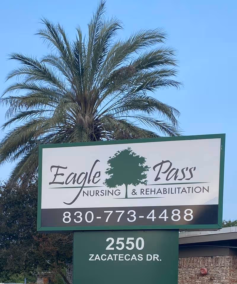 Outdoor sign for Eagle Pass Nursing & Rehabilitation with a phone number 830-773-4488 and address 2550 Zacatecas Dr. A large palm tree is visible behind the sign against a clear blue sky.