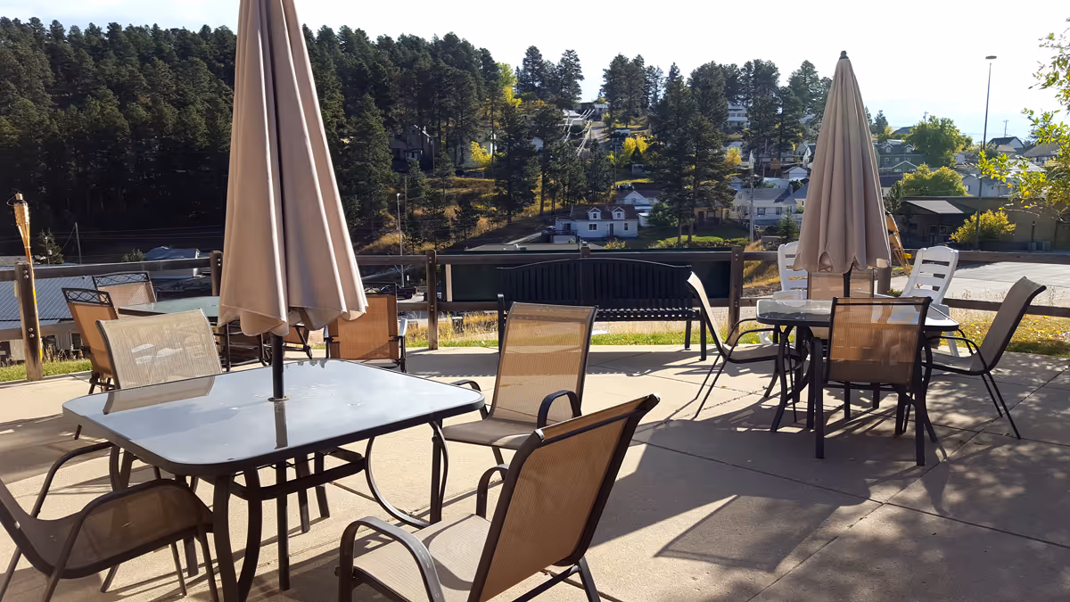 Outdoor patio area with glass-top tables and beige umbrellas, surrounded by mesh chairs. In the background, there are trees, houses, and a bench along a wooden fence under a clear sky.