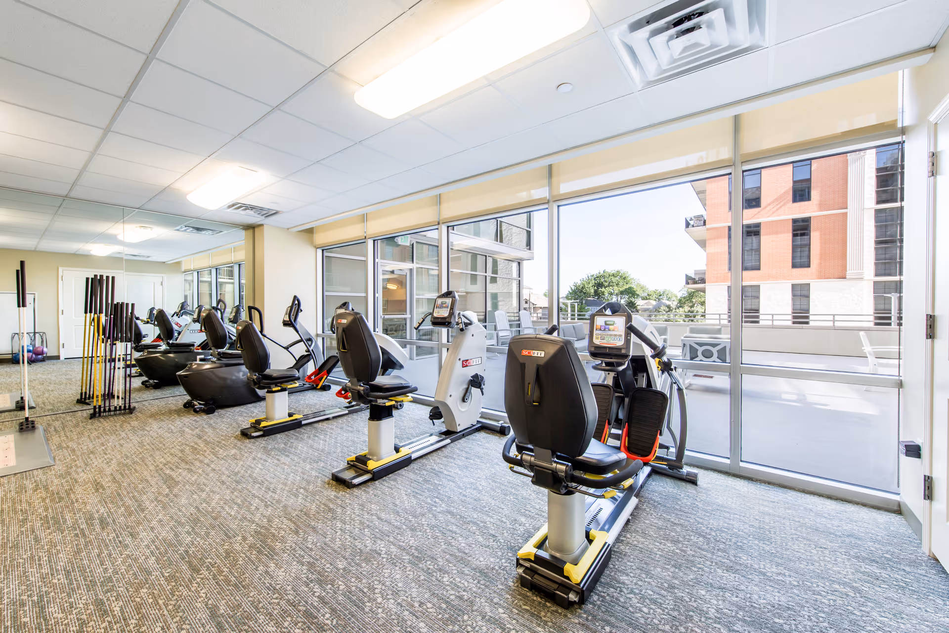 Bright fitness room with several exercise bikes and other workout equipment arranged in a row facing large windows that let in natural light and offer a view of an outdoor patio and nearby buildings.