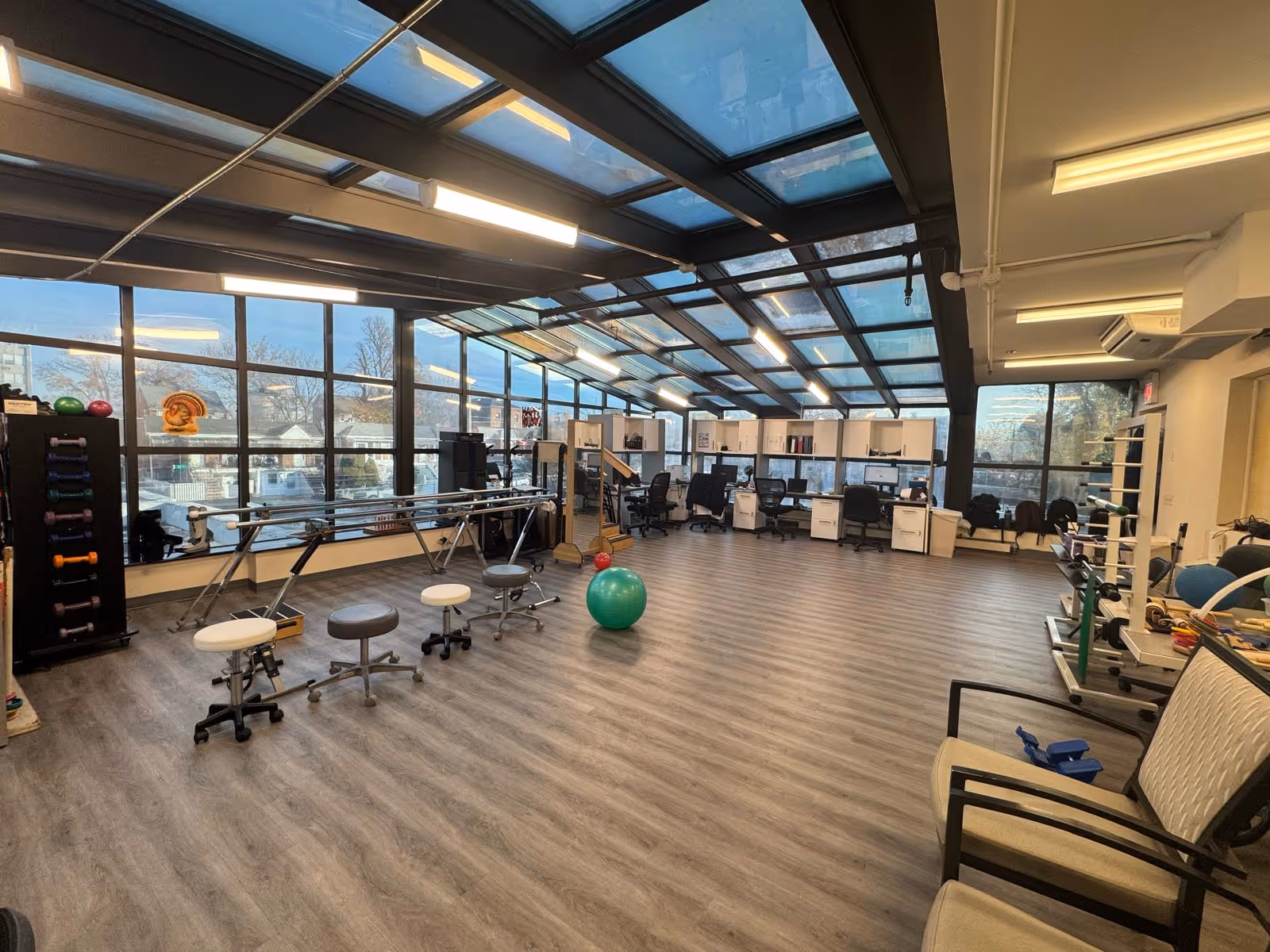 A spacious rehabilitation or therapy room with large windows and a glass ceiling allowing natural light. The room contains exercise equipment including stools, parallel bars, a green exercise ball, and various weights. There are desks and chairs along the far wall, likely for staff use. The floor is wood-patterned, and the room has a clean, organized appearance.