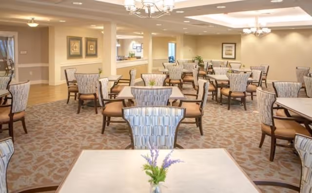 Spacious dining room with multiple square tables, patterned upholstered chairs, chandeliers, and small floral centerpieces.