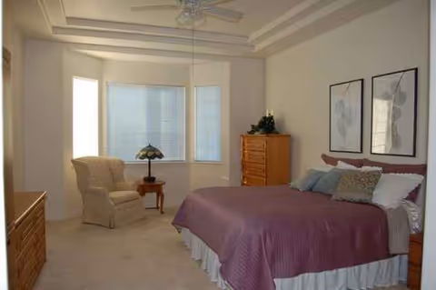 A cozy bedroom with a purple bedspread, dresser, chair, and soft natural light.