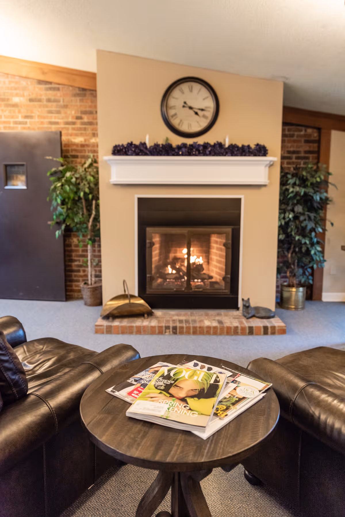 A cozy living room area with a lit fireplace surrounded by a brick hearth and beige walls. Above the fireplace is a white mantel decorated with purple garland and candles, and a round clock showing the time as 4:15. On either side of the fireplace are potted green plants. In the foreground, there is a round wooden table with magazines on it, flanked by two black leather armchairs.