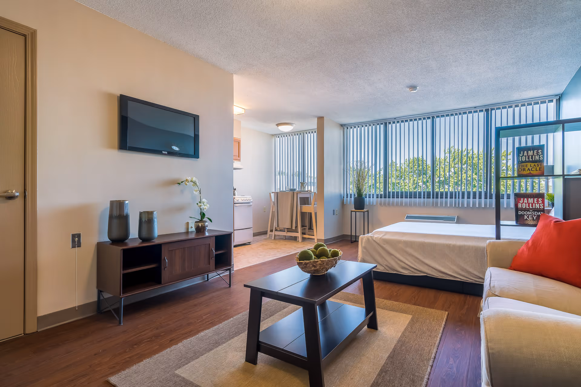 A bright and modern studio apartment with a bed, beige sofa with a red pillow, a dark wood coffee table with a bowl of green decorative balls, a TV mounted on the wall above a dark wood console table with decorative vases and a small plant. Large windows with vertical blinds let in natural light, and a small dining area with two chairs is visible near the kitchen area.