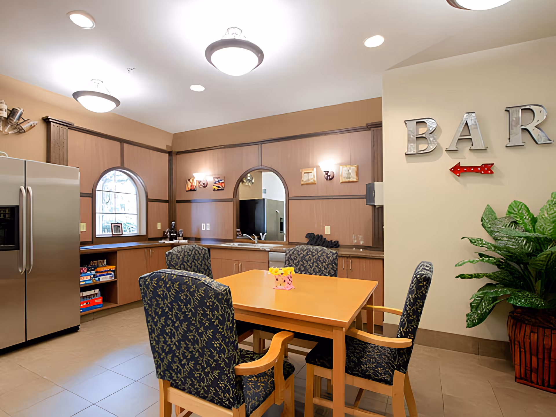 A cozy indoor bar area with a wooden table surrounded by four cushioned chairs with patterned upholstery. The room features a stainless steel refrigerator, wooden cabinets, a sink, and decorative wall lights. A large plant is placed in the corner, and metallic letters spelling 'BAR' with a red arrow are mounted on the wall.