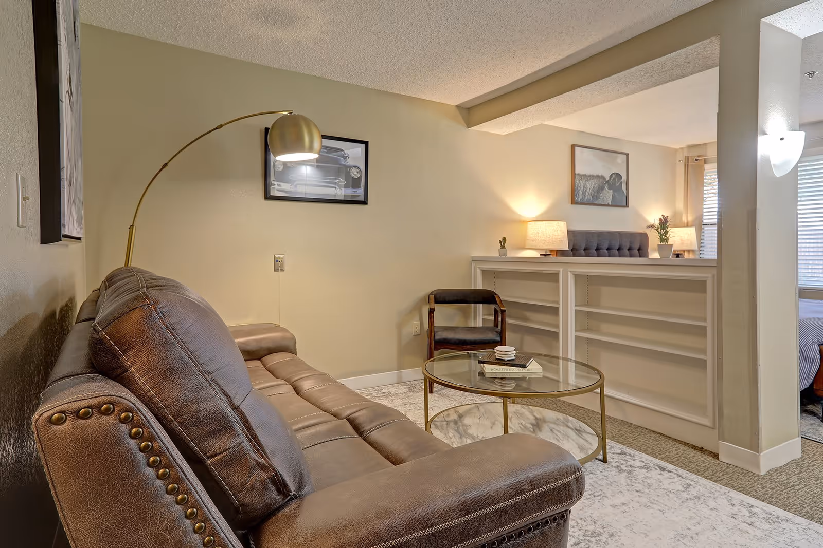 A cozy living room area featuring a brown leather sofa with nailhead trim, a round glass coffee table with a marble lower shelf, and a dark wooden chair. The room has beige walls adorned with framed pictures, a tall arc floor lamp, and a white built-in bookshelf with two table lamps and a small plant on top. Natural light filters in through a window with blinds in the background.