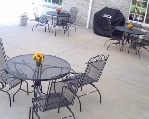 Outdoor patio area with multiple black metal tables and chairs. Each table has a small flower pot with yellow and orange flowers. There is a covered grill against a brick wall and windows in the background.