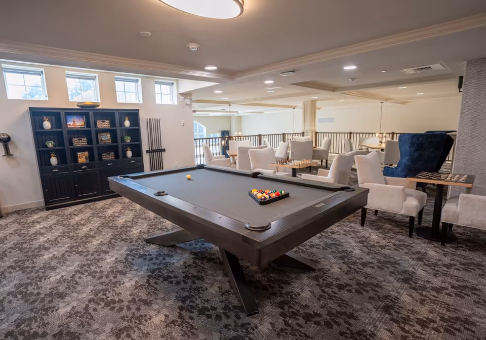 A bright communal lounge with a pool table centered on patterned carpet, surrounded by chairs and shelving.