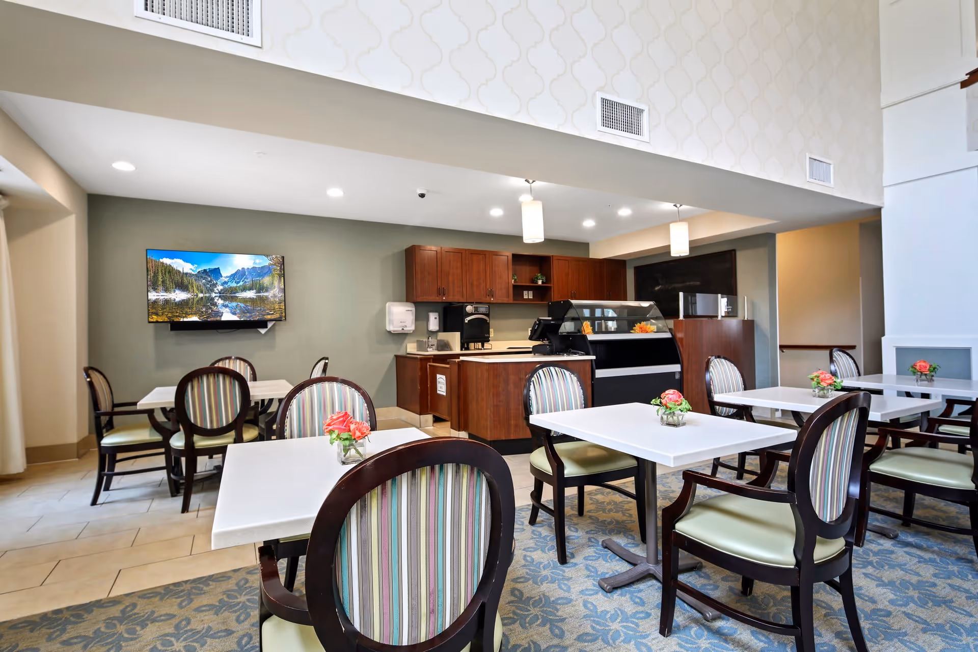 A bright dining area in a senior living facility featuring several white square tables with dark wooden chairs that have striped upholstery. Each table has a small vase with pink flowers. In the background, there is a counter with wooden cabinets, a coffee machine, and a display case. A flat-screen TV mounted on the wall shows a scenic nature image. The room has a high ceiling with recessed lighting and pendant lights.