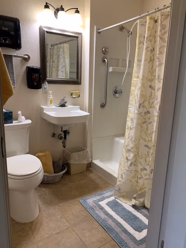 A clean and well-lit bathroom with a toilet, a small wall-mounted sink with a soap dispenser, a mirror above the sink, and a shower with a floral-patterned curtain. There is a towel hanging on a rack, a waste bin, and a small basket with towels on the floor. The floor is tiled and there is a blue and white striped bath mat in front of the shower.