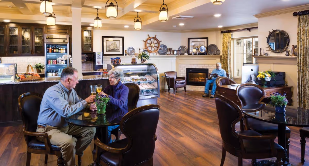 A cozy common area in Canterbury Park Independent Living Community featuring a couple seated at a round glass table with a small potted plant, enjoying drinks. The room has wooden flooring, leather chairs, a fireplace, and a display case with baked goods. Another person is seated in the background reading near a window with curtains. The space is warmly lit with hanging lights and decorated with nautical-themed items and framed pictures.