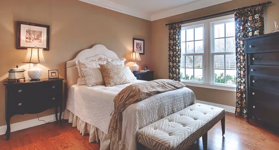 A cozy bedroom with a neatly made bed featuring multiple pillows and a beige quilt. There are two dark wooden nightstands on either side of the bed, each with a lamp and decorative items. A cushioned bench is placed at the foot of the bed. The room has hardwood floors, beige walls, and a large window with floral curtains allowing natural light to enter.