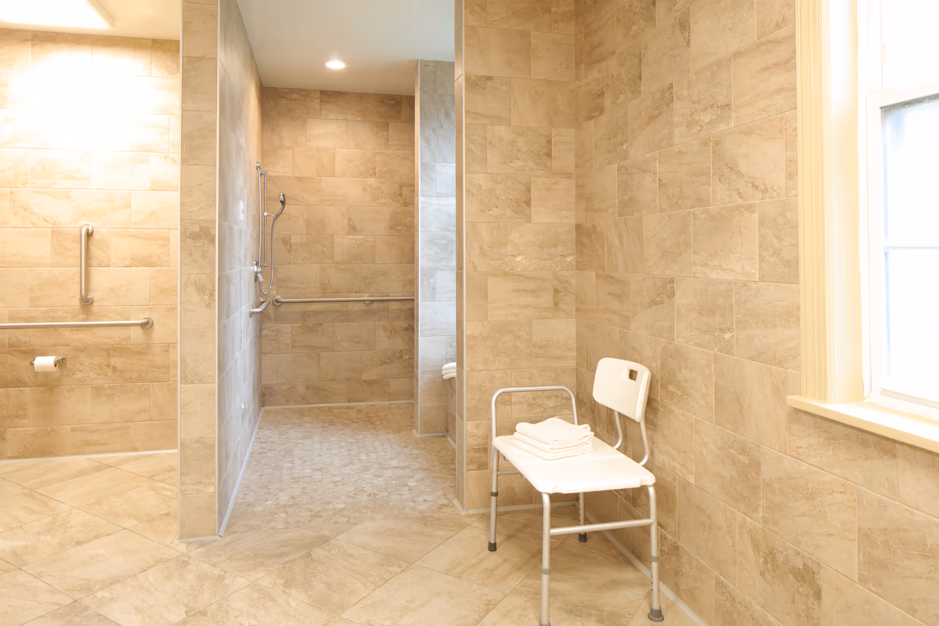 A spacious bathroom with beige tiled walls and floors, featuring a walk-in shower with grab bars and a handheld showerhead. A white shower chair with folded towels is placed near a window that allows natural light into the room.