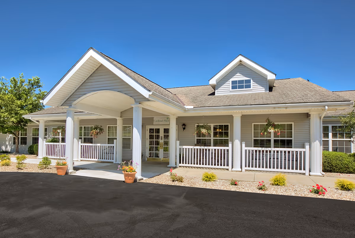 Exterior view of Cardinal Place senior living facility showing a single-story building with a covered entrance supported by white columns, a porch with white railings, hanging flower baskets, and a clear blue sky above.