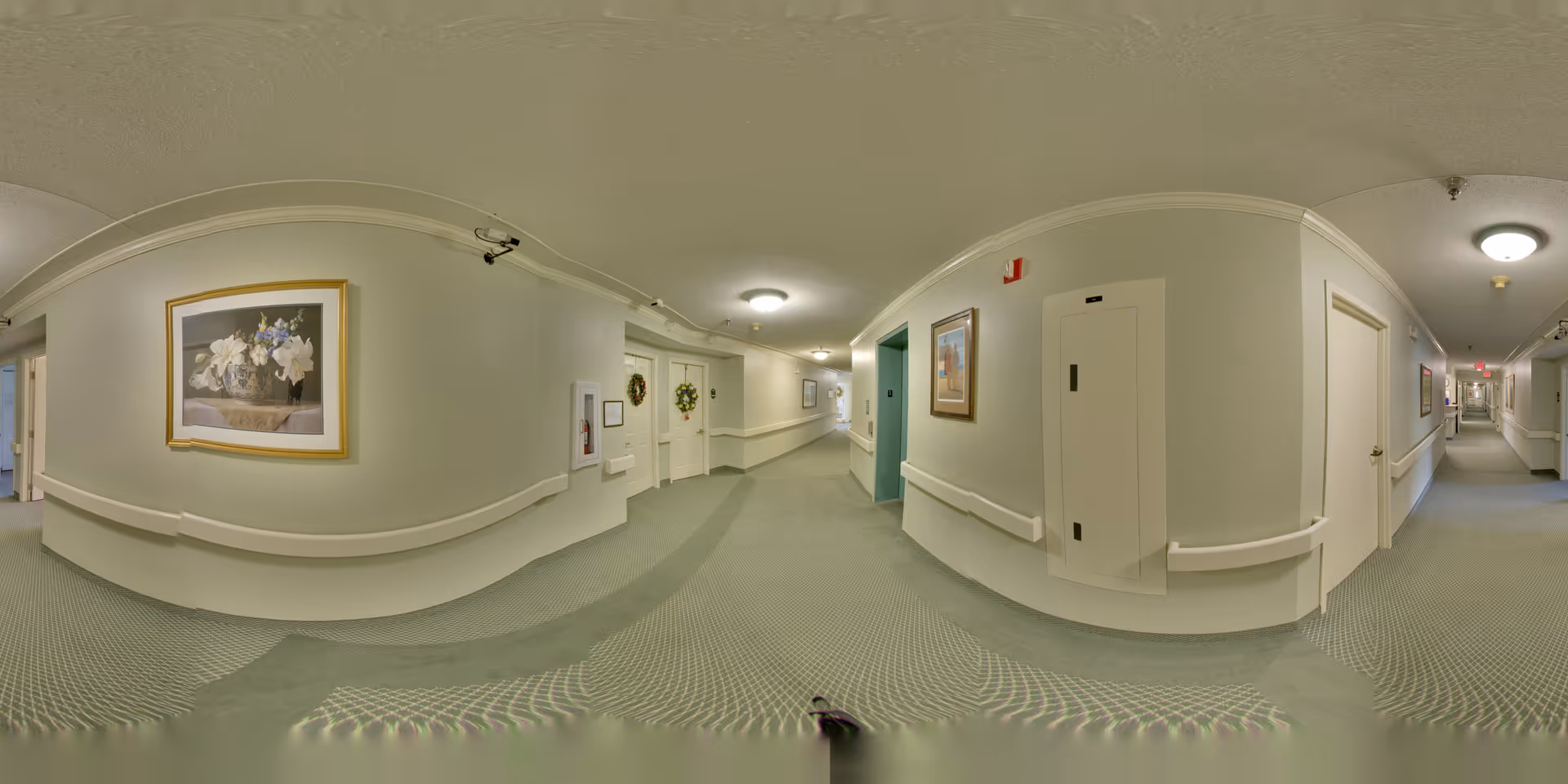 A long, well-lit hallway in a senior living facility with light green carpeting and cream-colored walls. The hallway features handrails on both sides, framed floral artwork, and several doors, some decorated with wreaths. Ceiling lights illuminate the corridor, and there is a fire extinguisher cabinet mounted on the wall.