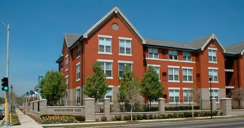 Exterior view of a three-story red brick senior living facility named Westminster Place with multiple windows, trees, and a stone fence along the sidewalk under a clear blue sky.