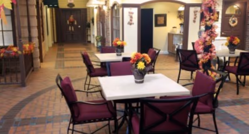 Indoor seating area with square tables and purple cushioned chairs arranged on a tiled floor. Each table has a vase with colorful flowers. The space is decorated with autumn-themed garlands and wreaths, and there are doorways and archways leading to other rooms in the background.