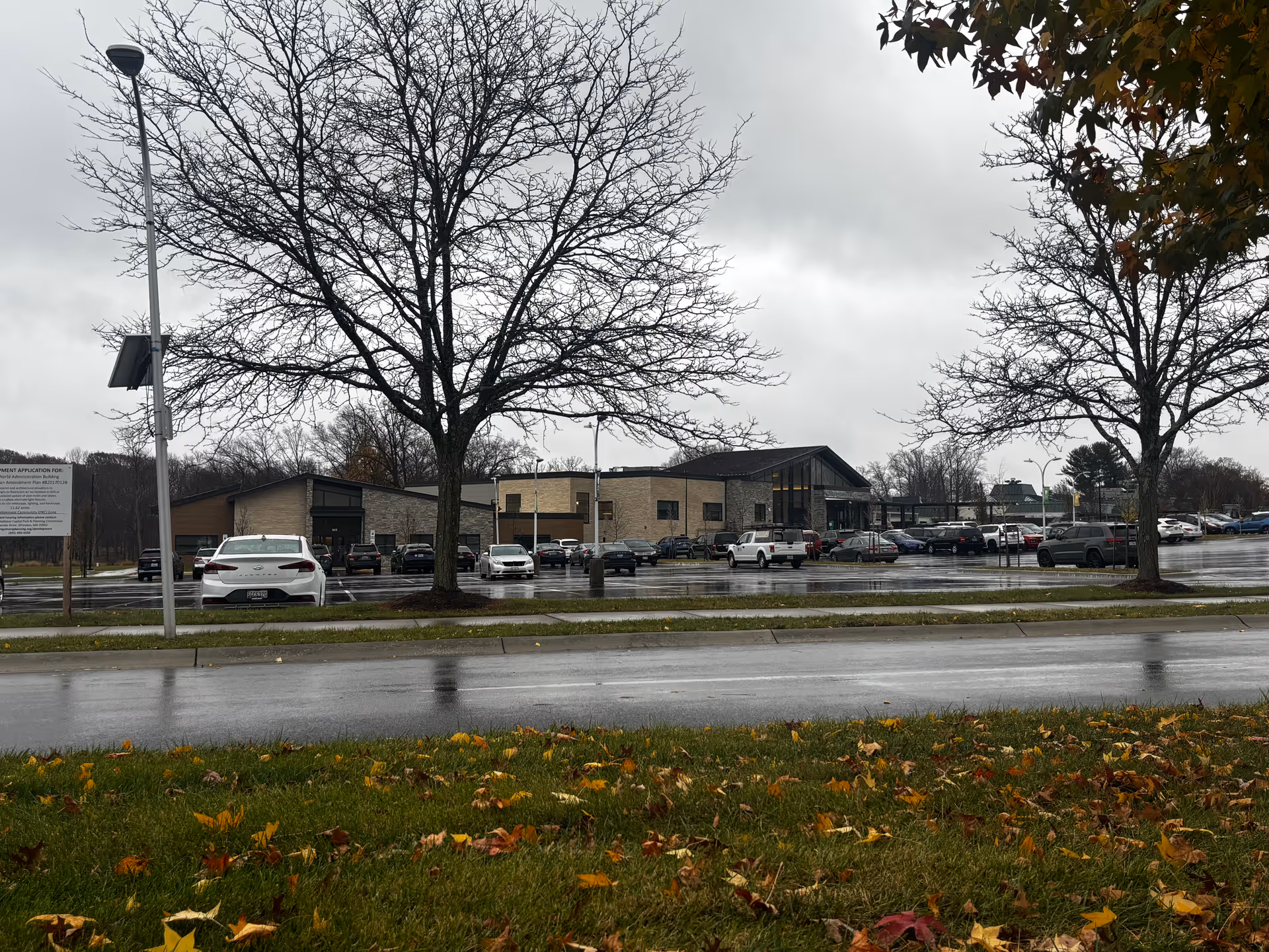Exterior view of a low-rise brick senior living building and parking lot seen across a wet road with bare trees and fallen leaves under an overcast sky.
